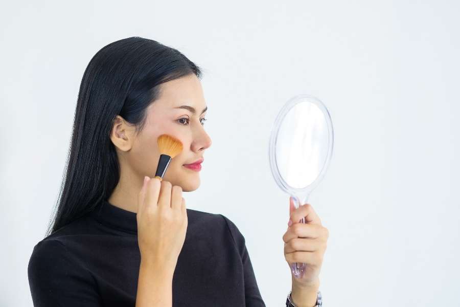 Une jeune femme aux cheveux noirs applique du blush avec un pinceau doré en se regardant dans un miroir à main, maîtrisant les techniques de maquillage de base.