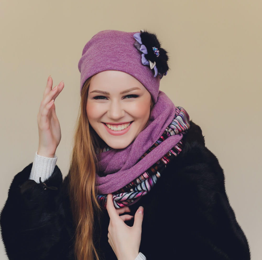 emme souriante portant un bonnet et l'un de nos foulards et écharpes de créateur en laine mauve avec des détails rayés, assortis à un manteau en fourrure noire.