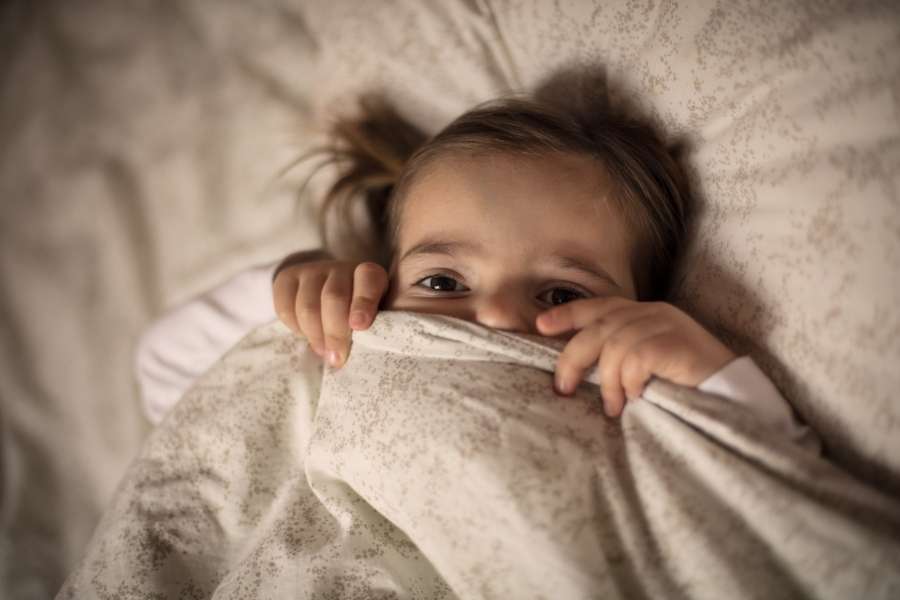 Un enfant dont les yeux écarquillés dépassent de la couette dans une ambiance sombre, capturant l'inquiétude quand un enfant se réveille la nuit.