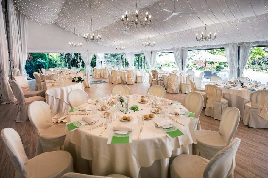 Intérieur d'un chapiteau de réception élégant avec des tables rondes nappées de blanc, des lustres et des guirlandes lumineuses sous un plafond drapé.