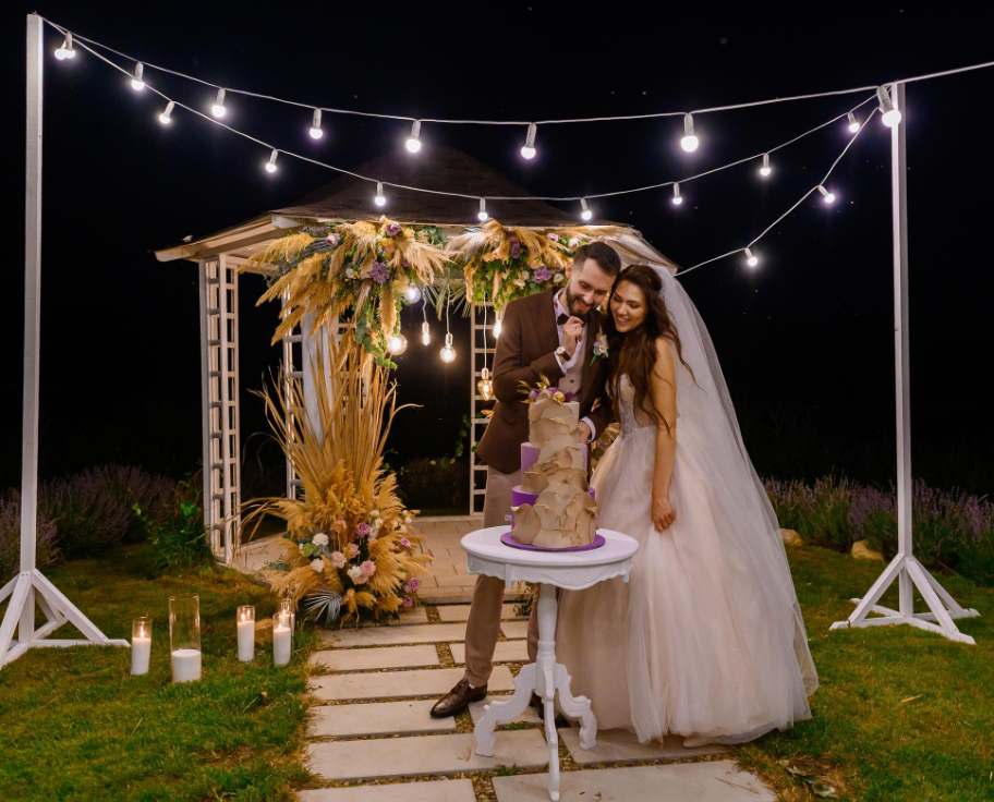 Mariés coupant leur gâteau sous des guirlandes et lumieres pour deco mariage nuit devant une gloriette fleurie.