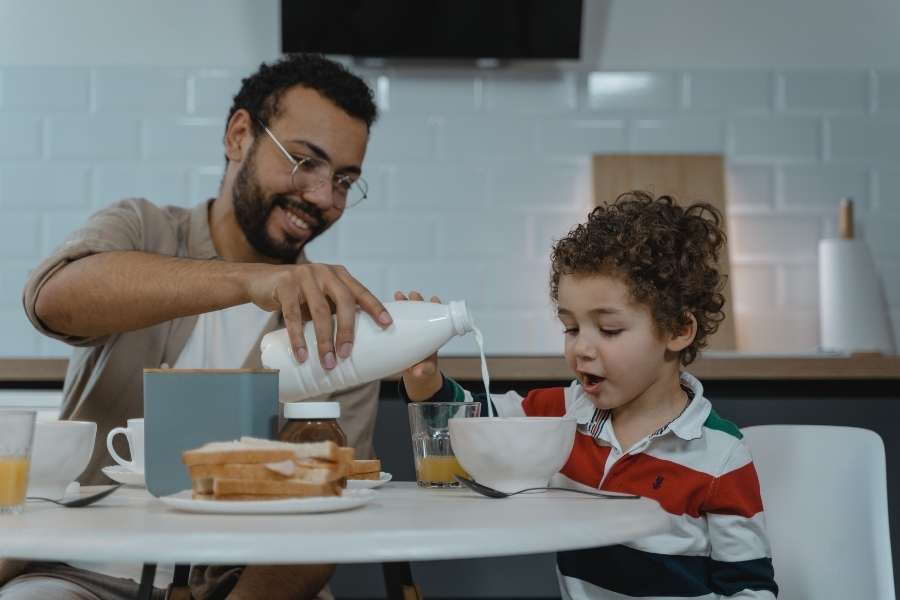 Un père verse du lait dans un bol pour son jeune enfant à table, illustrant les moments de partage lors de la diversification menée par l'enfant.